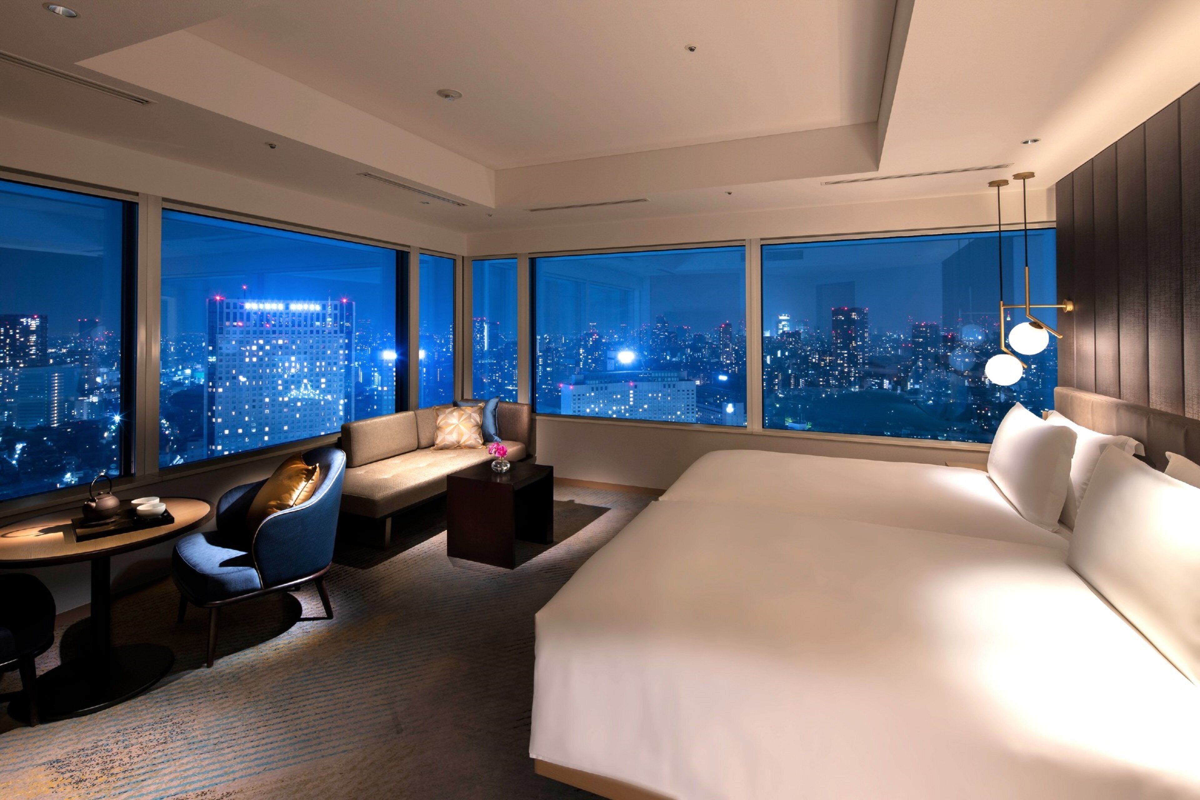 Une chambre d’hôtel avec un grand lit, un coin salon comprenant un canapé et un fauteuil, une petite table et une vue sur le paysage urbain la nuit.