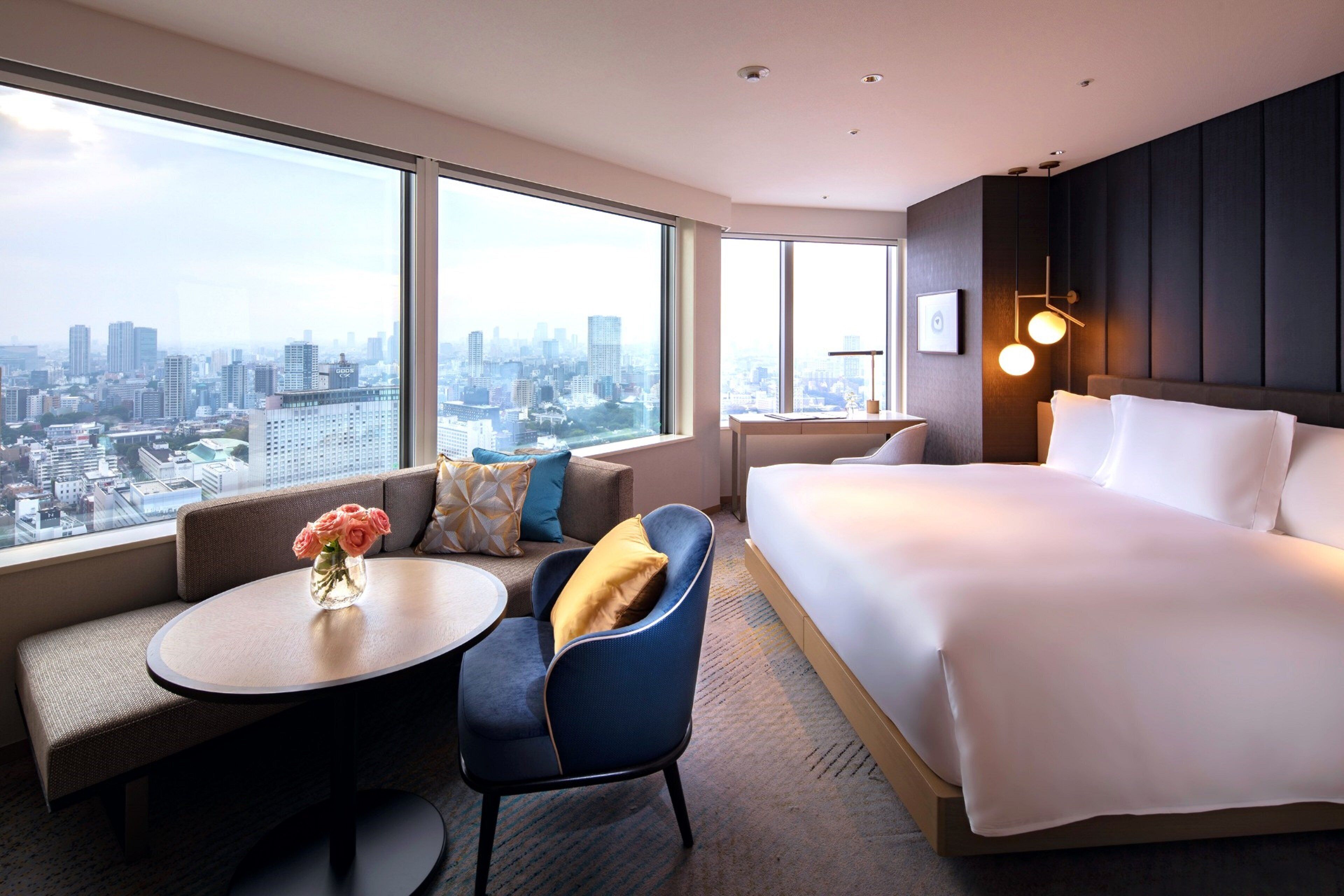 A hotel room with a large bed, a seating area with a table, and a view of a cityscape through large windows.