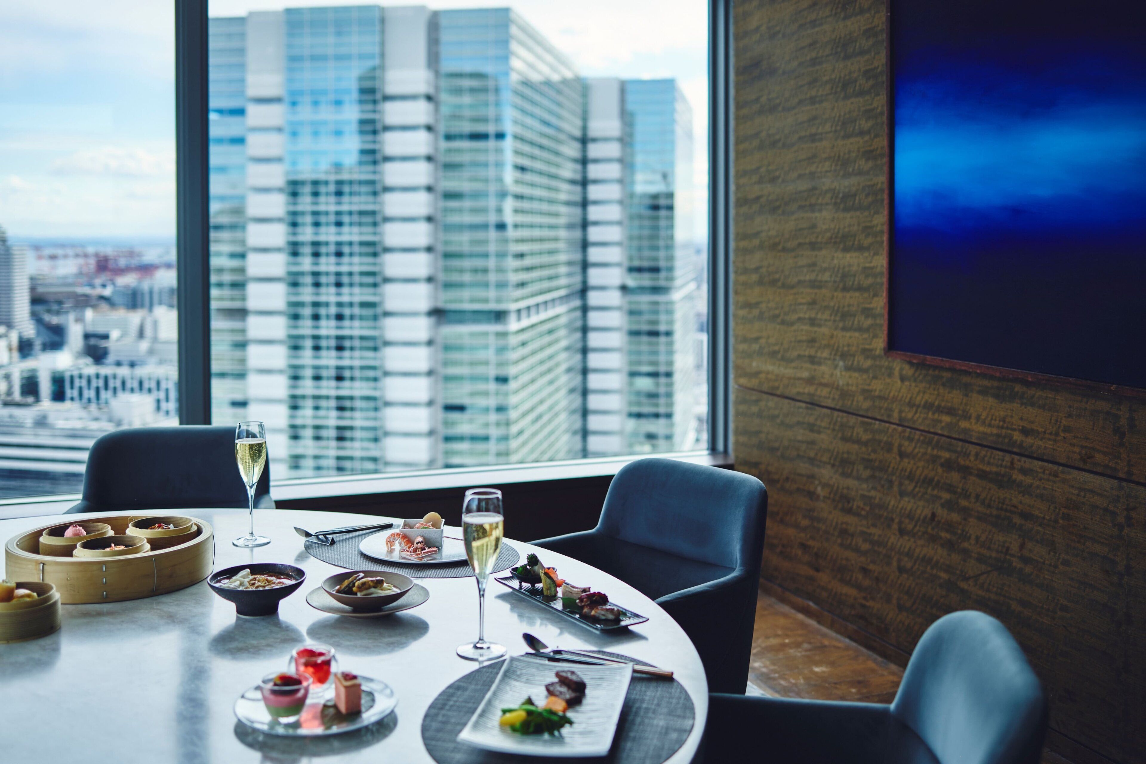 A dining table with various dishes and a glass of wine, set against a cityscape view.