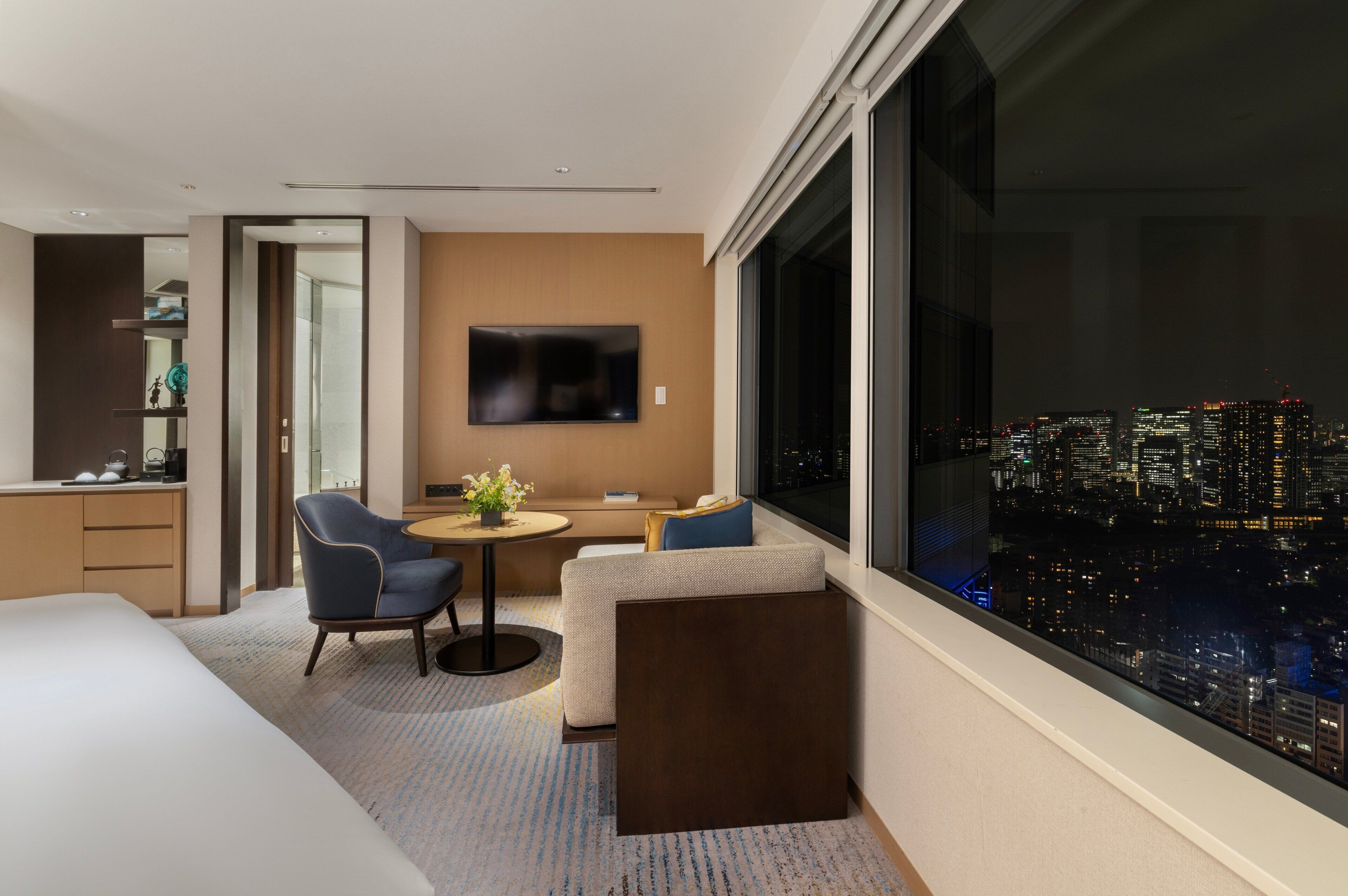 Une chambre d’hôtel moderne équipée d’un lit, d’un bureau, d’une chaise, d’un canapé, d’une petite table avec un vase de fleurs et d’un téléviseur fixé au mur.