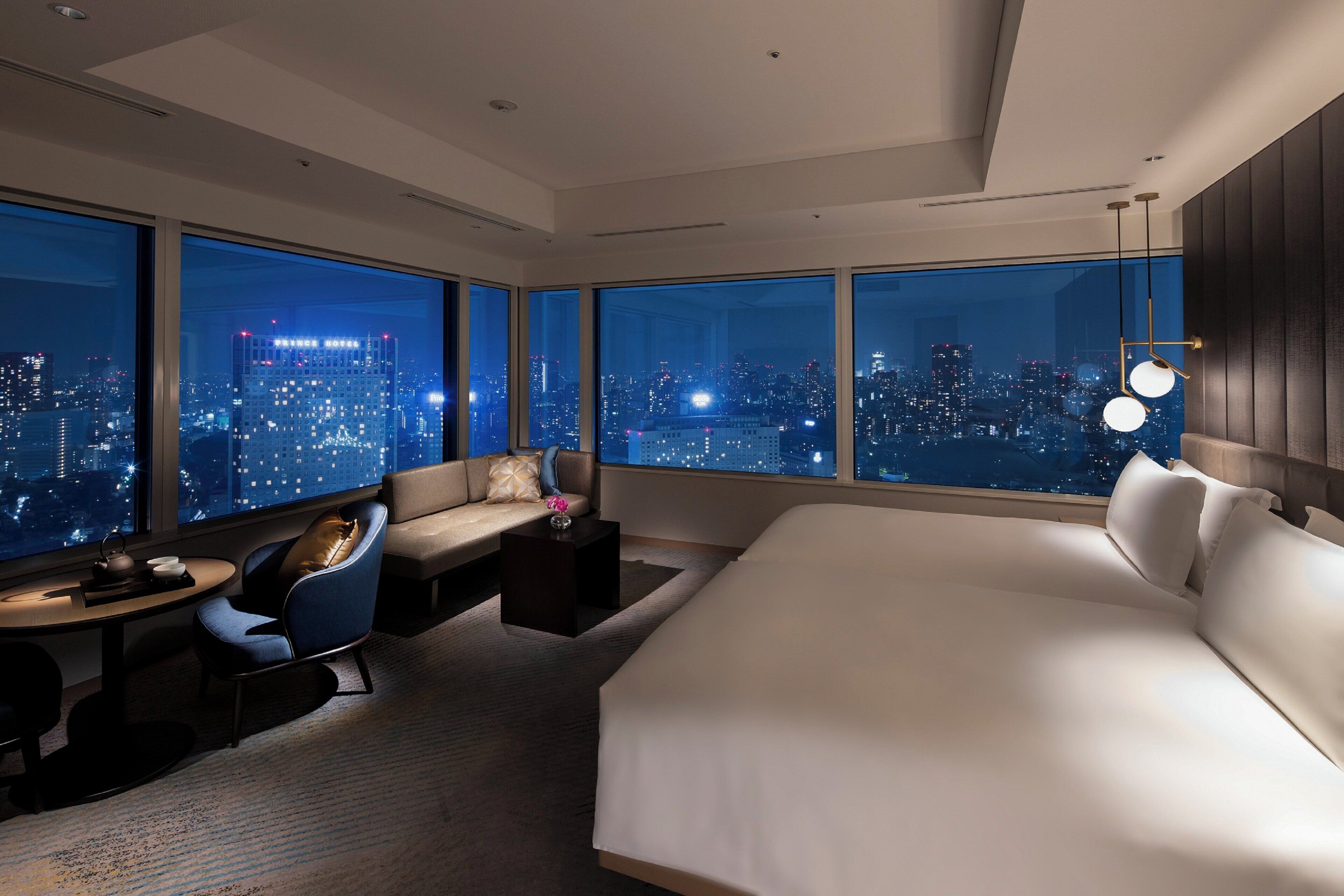 Une chambre d’hôtel moderne avec un grand lit, un coin salon comprenant un canapé et un fauteuil, une petite table et une vue sur la ville la nuit.