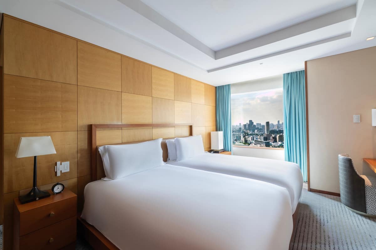 Une chambre d’hôtel avec deux lits, une tête de lit en bois, une table de chevet avec une lampe, une vue sur la ville et une fenêtre avec des rideaux.