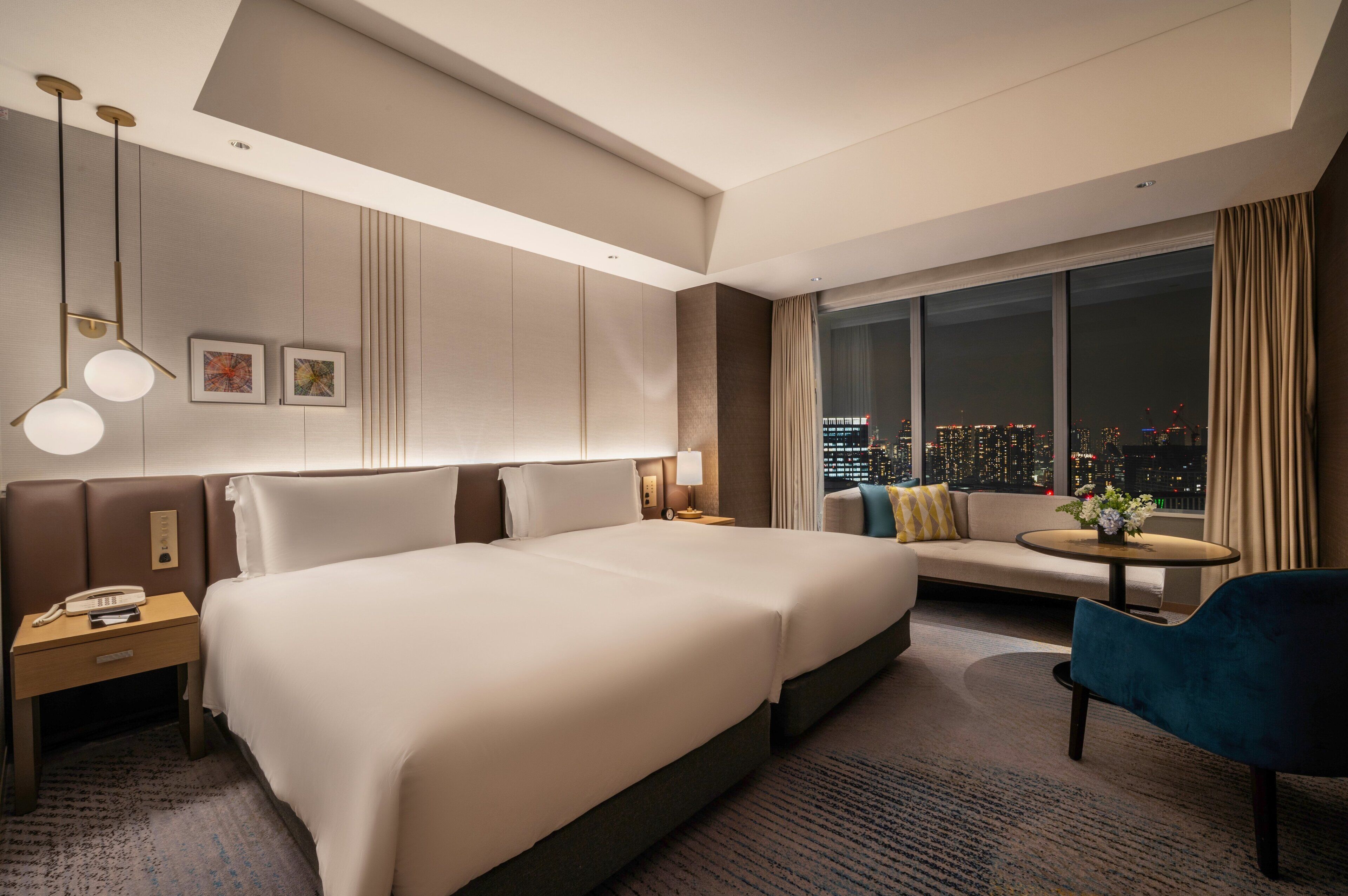 A hotel room with a large bed, a sofa, a small table with flowers, a nightstand with a phone, and a view of the city at night.