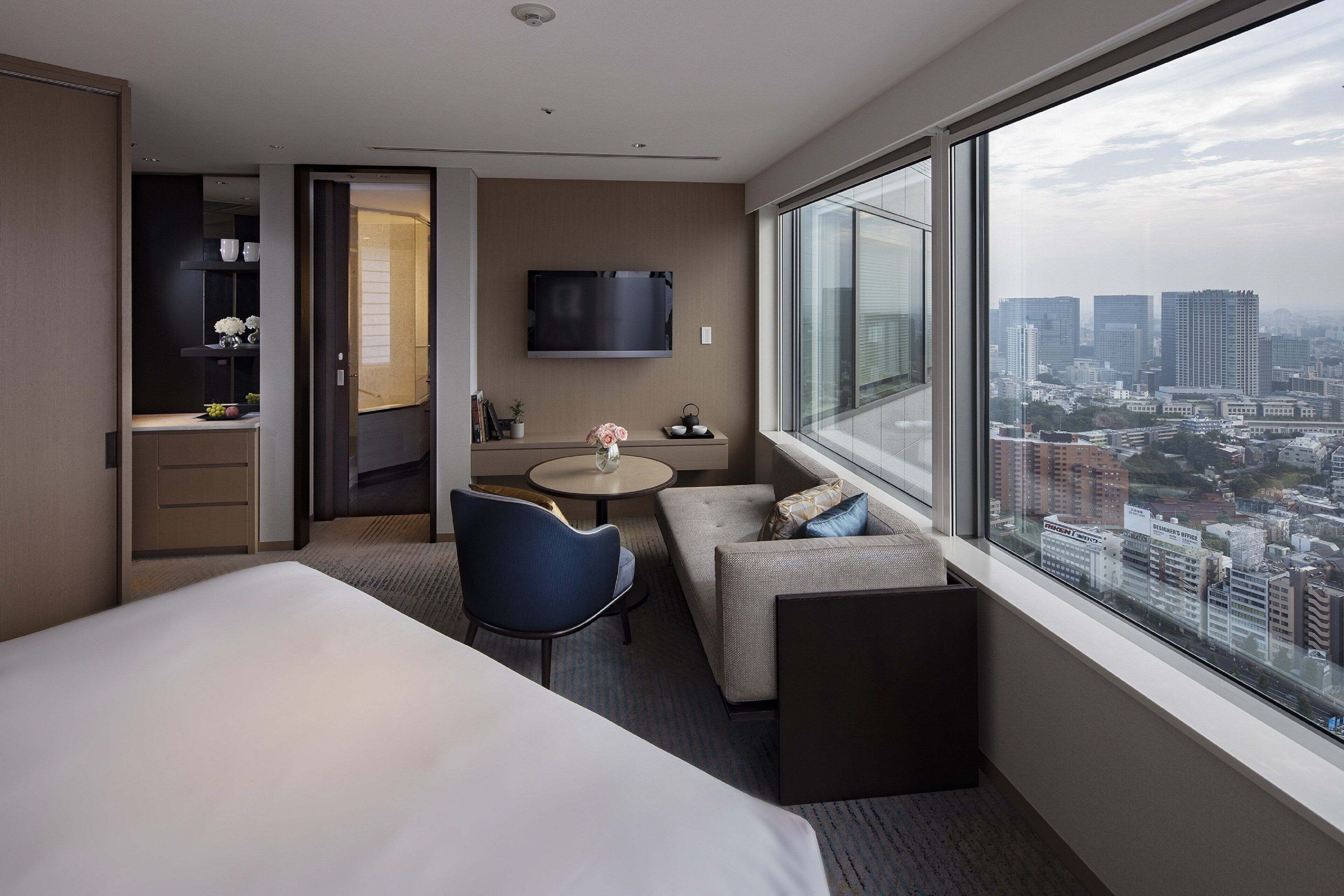 A modern hotel room with a large bed, a seating area with a table, a TV mounted on the wall, and a city view through large windows.