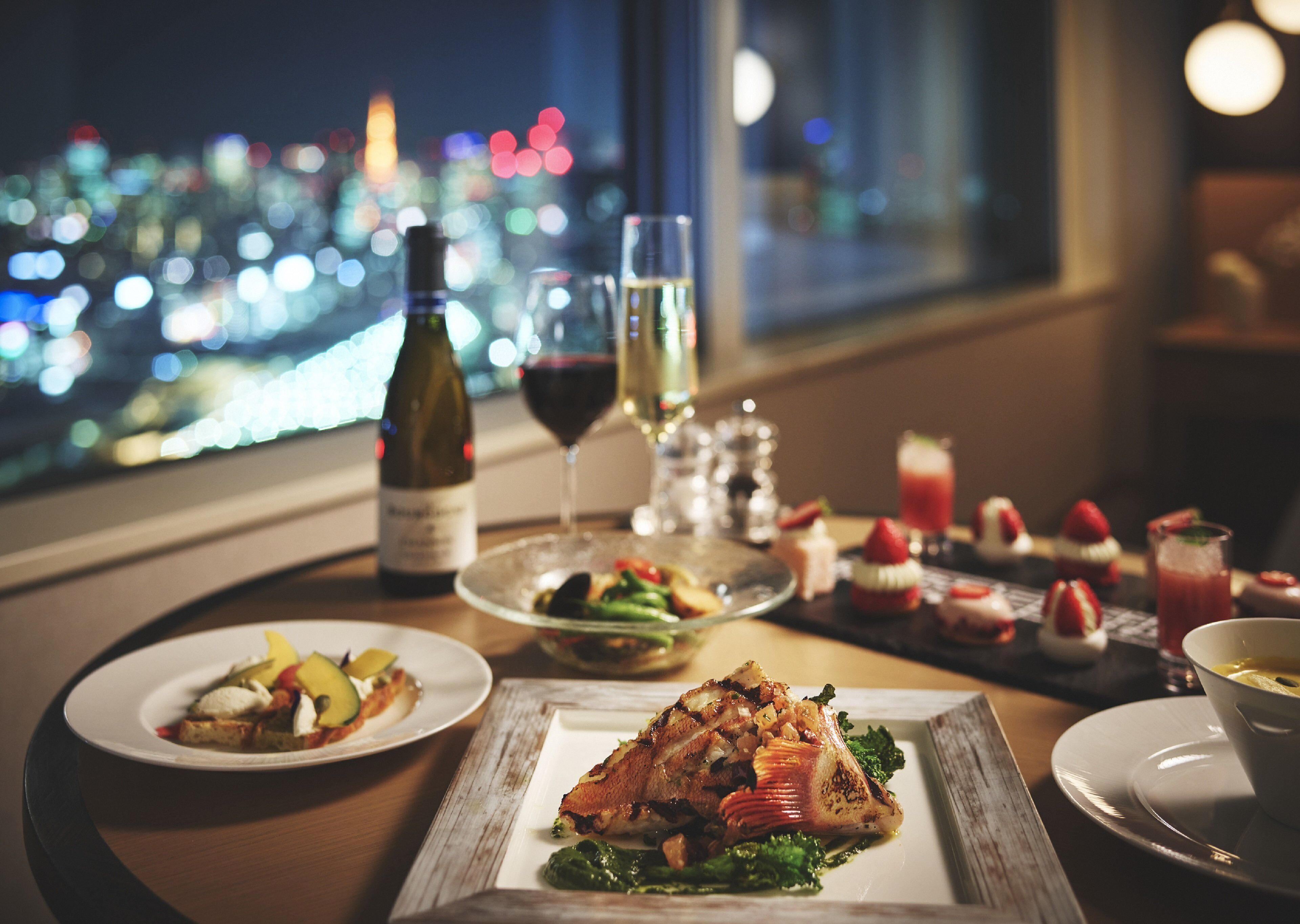 A dining table with plates of food, a bottle of wine, and glasses of wine, set against a cityscape view.
