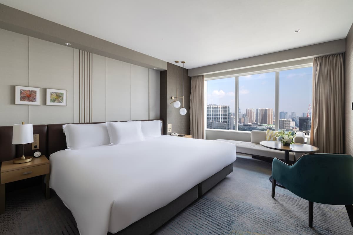 Une chambre d’hôtel moderne avec un grand lit, une table de chevet avec une lampe, une petite table avec un vase, un canapé et une vue sur le panorama de la ville.