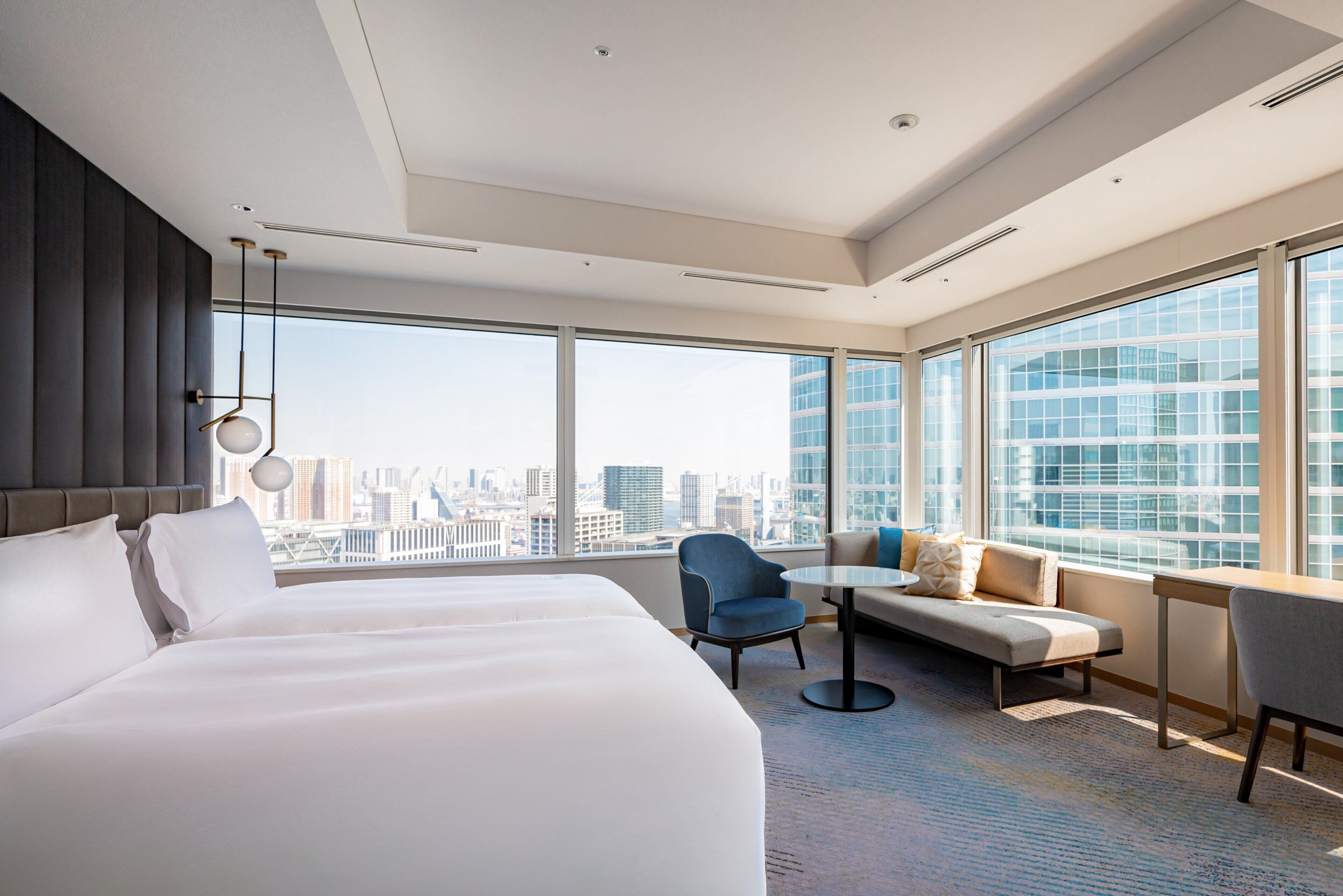 A modern hotel room with a large bed, a seating area with a sofa and chairs, a small table, and a large window offering a city view.