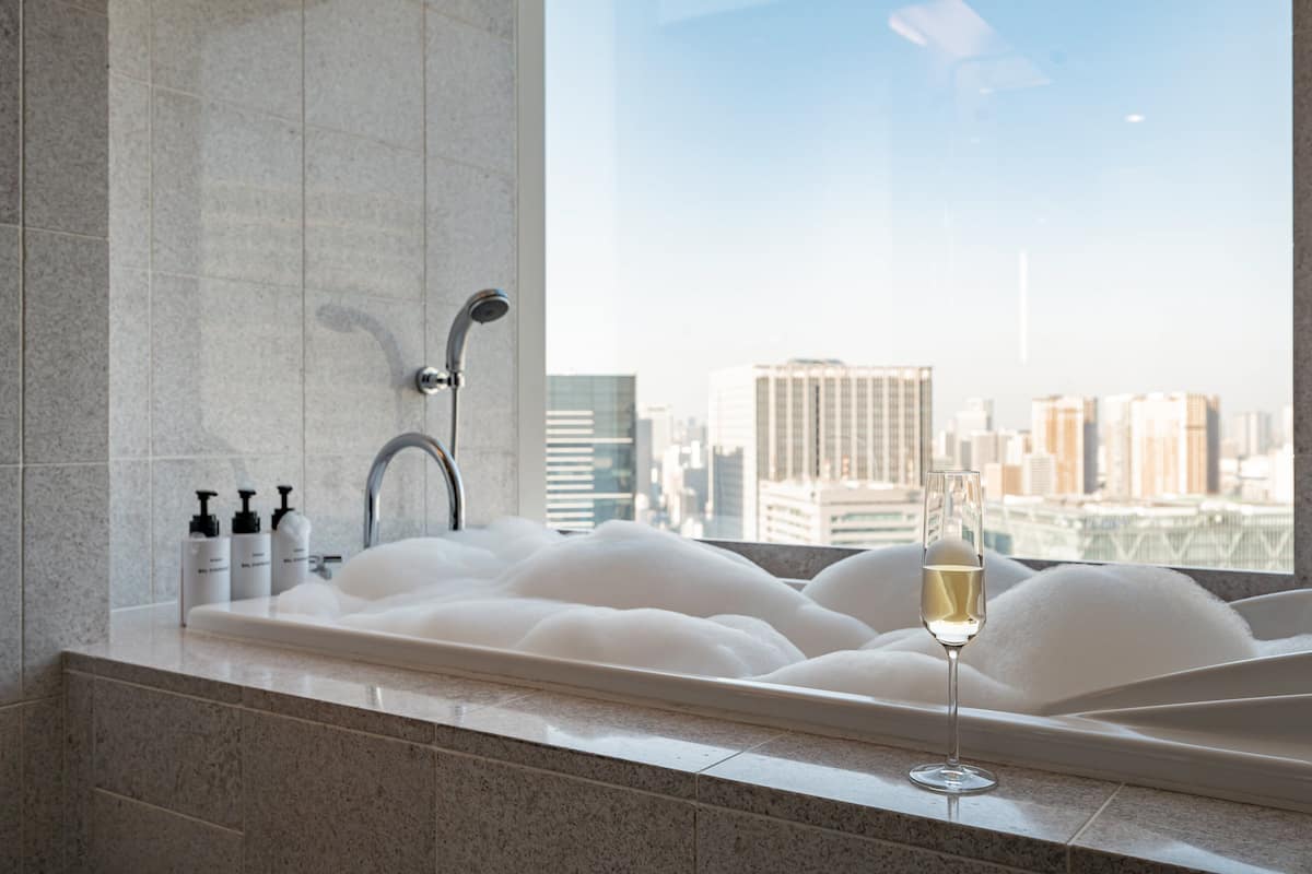 Une salle de bain moderne avec une grande baignoire remplie de mousse, une coupe de champagne et une vue sur le paysage urbain, visible à travers une grande fenêtre.