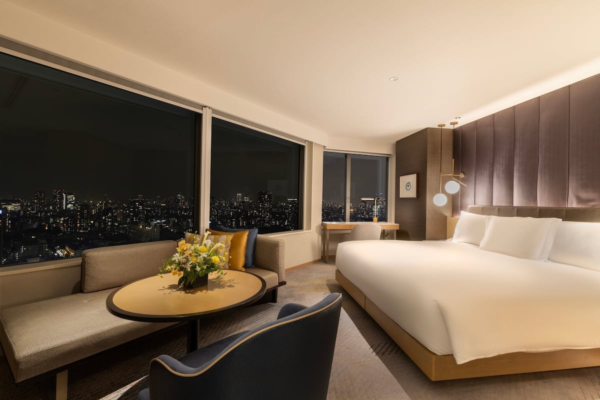 Une chambre d’hôtel moderne avec un grand lit, un canapé, une table ronde agrémentée de fleurs et une vue sur la ville à travers de grandes fenêtres.