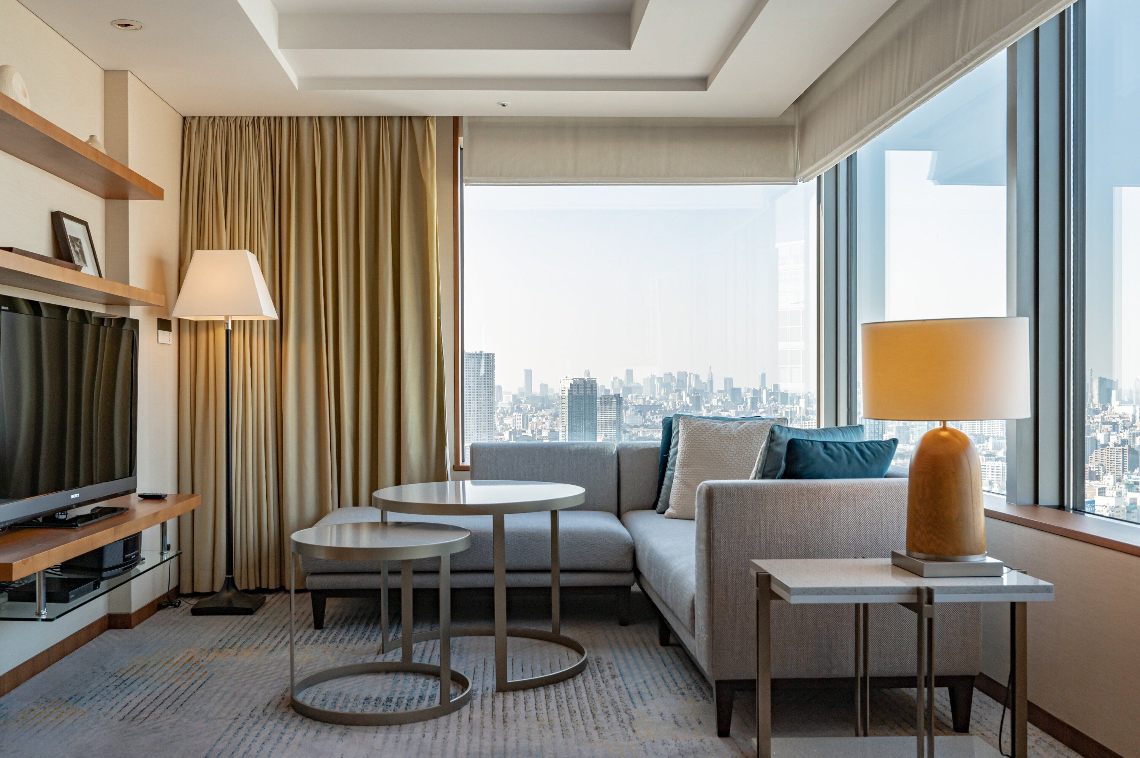 A modern hotel room with a city view, a sofa, two small tables, a TV, and a lamp.