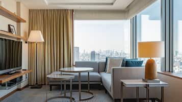 A modern hotel room with a city view, a sofa, two small tables, a TV, and a lamp.