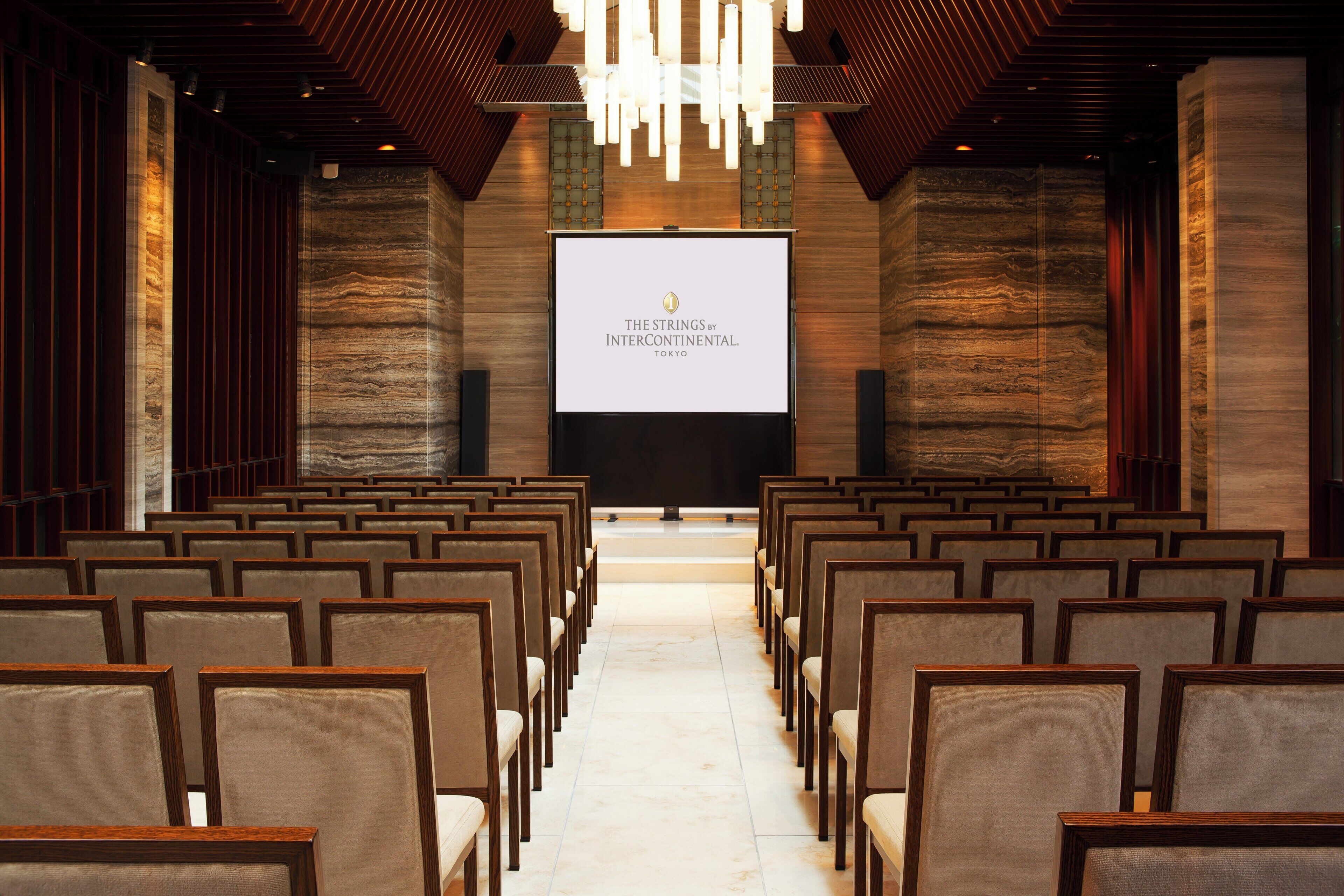 An auditorium with rows of chairs, a large screen displaying 'THE STRINGS BY INTERCONTINENTAL TOKYO', and a modern chandelier.