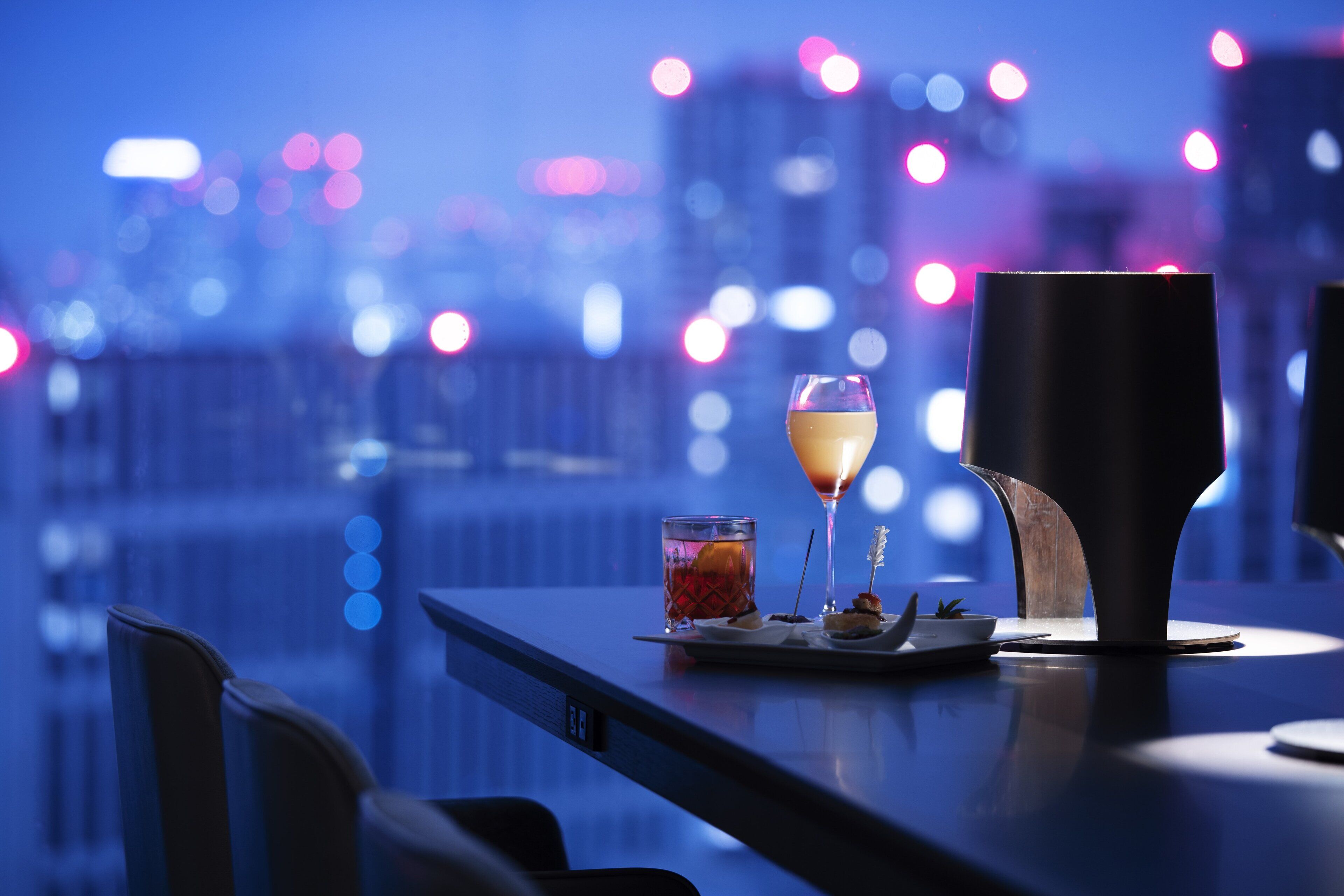 A table with a glass of drink, a dessert, and a candle in a dimly lit room with a cityscape view.