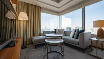 A modern hotel room with a sofa, two round tables, a TV, and large windows offering a city view.