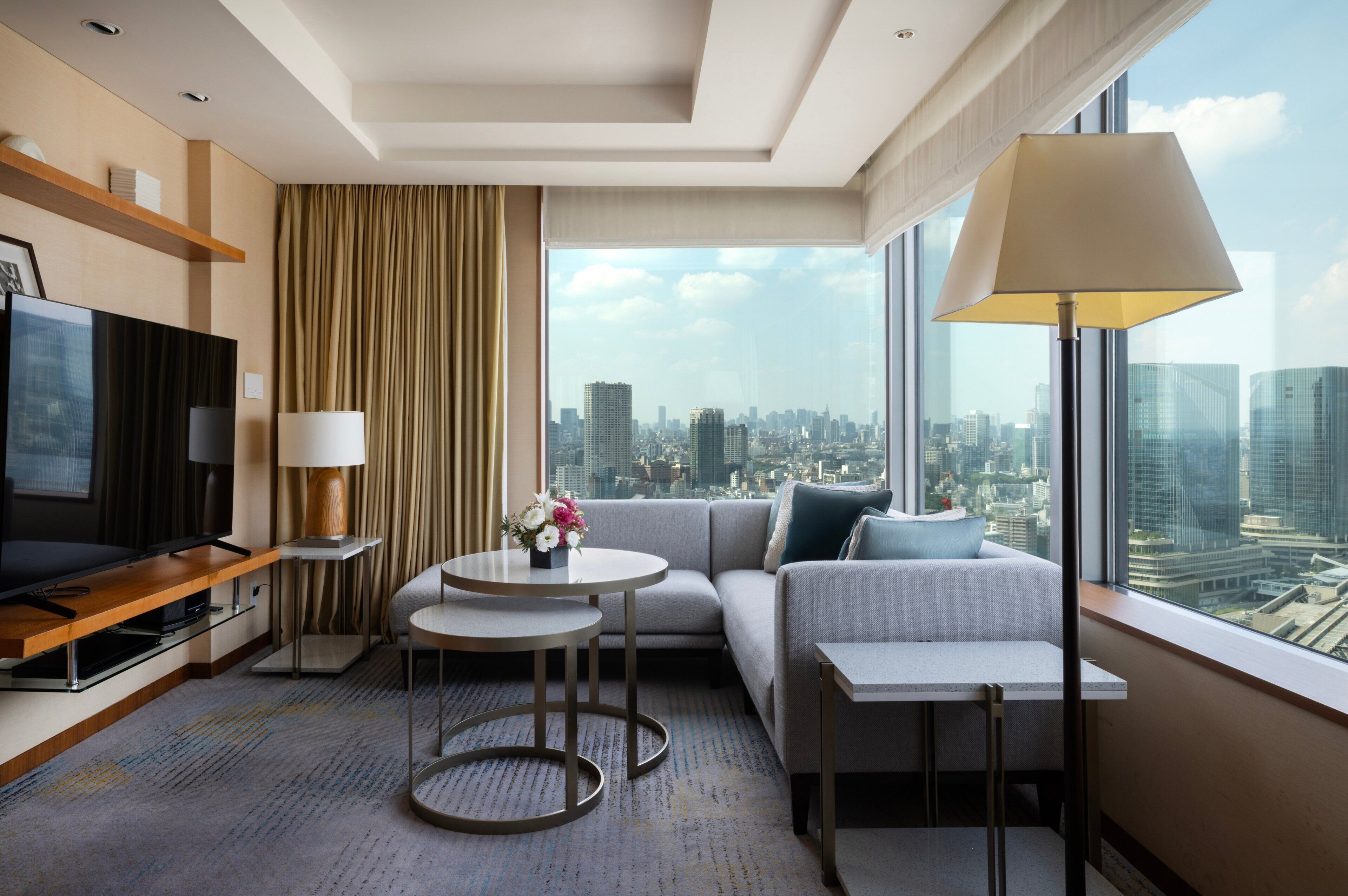 A modern hotel room with a large window, a sofa, a TV, and a small table with a lamp.