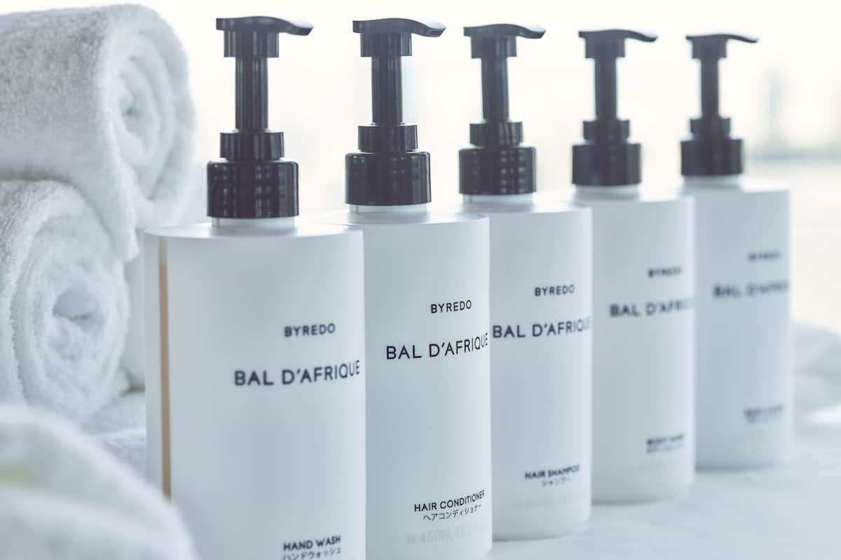 Une rangée de flacons blancs avec des pompes noires, étiquetés « BYREDO BAL D’AFRIQUE », et une serviette blanche roulée en arrière-plan.