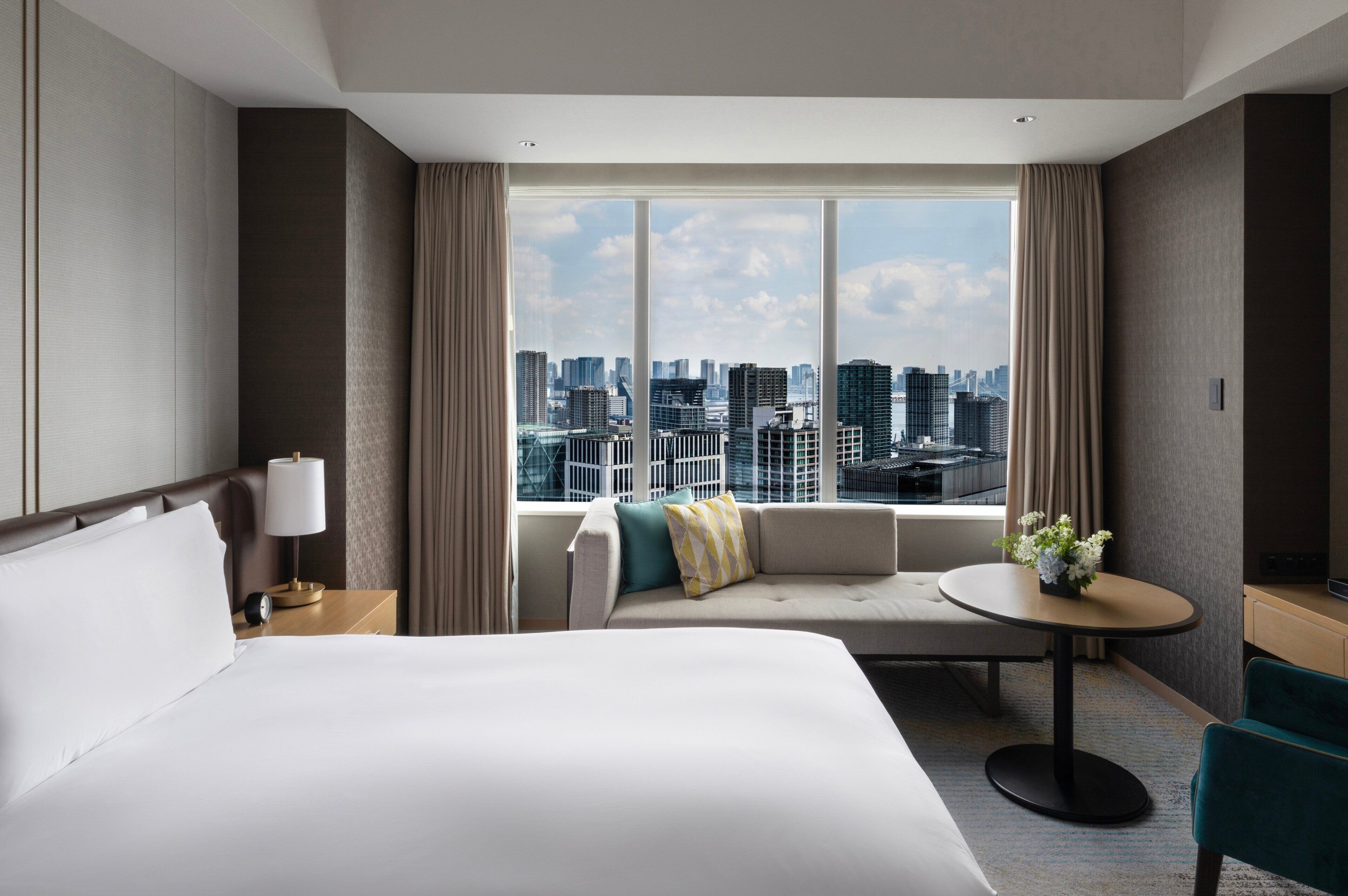 Habitación de hotel con una cama grande, un sofá, una mesita y vistas a un paisaje urbano a través de amplios ventanales.