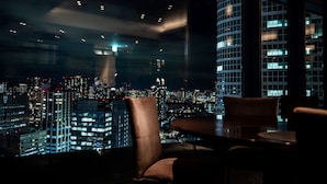 A high-rise hotel room with a panoramic city view, a large window, and a dining area with a table and chairs.