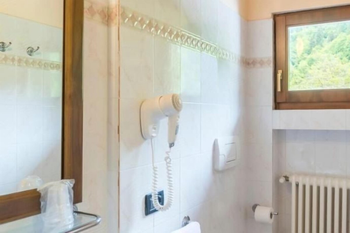 A bathroom with a hairdryer mounted on the wall, a window with a view of greenery, and a radiator.