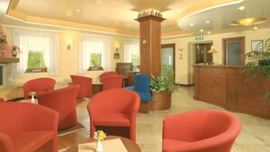 A hotel lobby with red armchairs, a wooden reception desk, and a large potted plant. - Hotel Conca Verde (Zone)