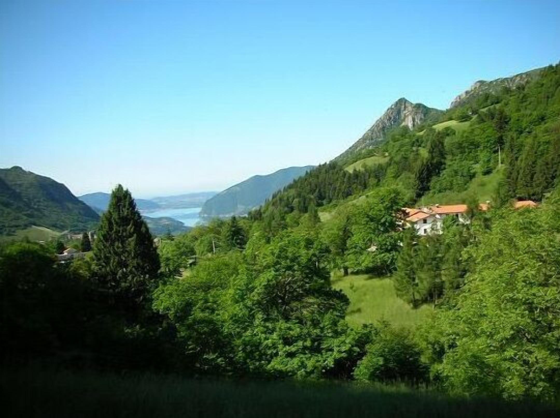 Eine gebirgige Landschaft mit einem Haus, umgeben von dichtem Grün und blauem Himmel.