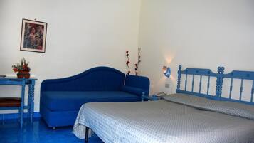 A bedroom with a blue sofa, a bed with a blue headboard, a small table with a vase, and a wall-mounted lamp.