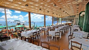 Un espace de restauration intérieur avec des tables dressées, de grandes fenêtres offrant une vue sur la mer et les bateaux.