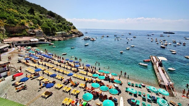 Une plage dotée de nombreuses chaises longues et de parasols, une jetée s’avançant dans la mer, et des bateaux ancrés à proximité.