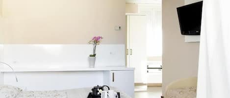 Une chambre d’hôtel avec un lit, une table de chevet ornée d’un vase de fleurs, un plateau contenant divers objets, un téléviseur fixé au mur et une porte donnant sur une autre pièce.