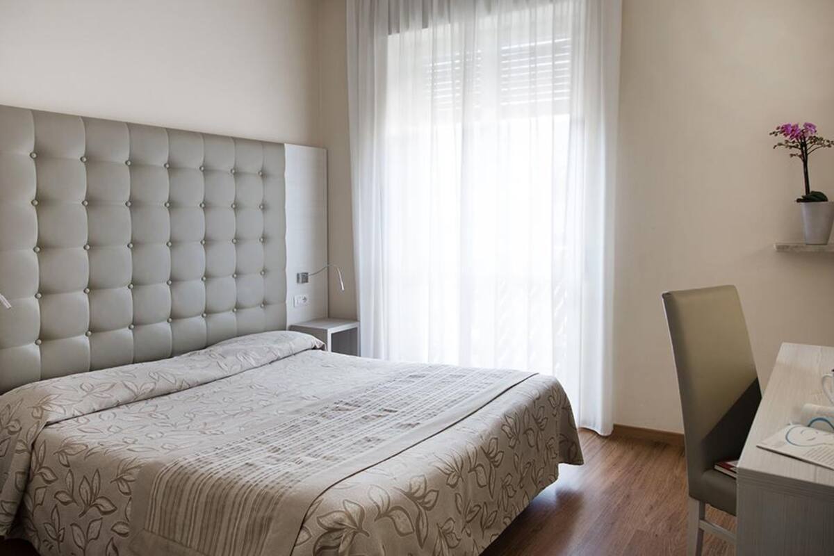Un dormitorio con un cabecero capitonado, una cama con colcha de estampado floral, una mesita de noche, una silla y una ventana con cortinas traslúcidas.