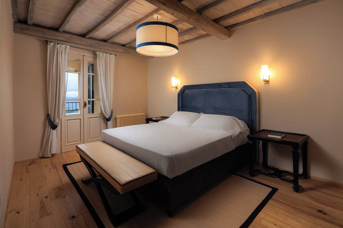 Ein Schlafzimmer mit einem Bett, Nachttischen, einer Bank, einem Fenster mit Vorhängen und einer Deckenleuchte.