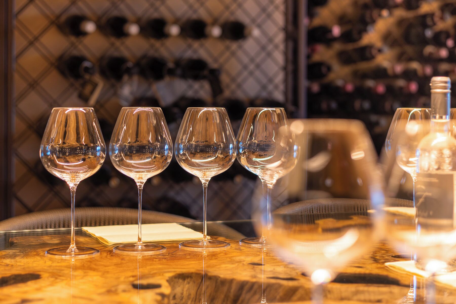 A row of empty wine glasses on a table with a wine rack in the background.