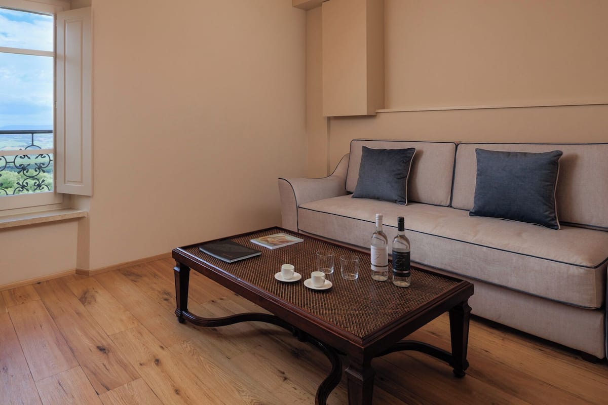 Ein Zimmer mit Holztisch, einer Couch mit Kissen, einem Fenster mit Blick und einer Flasche auf dem Tisch.