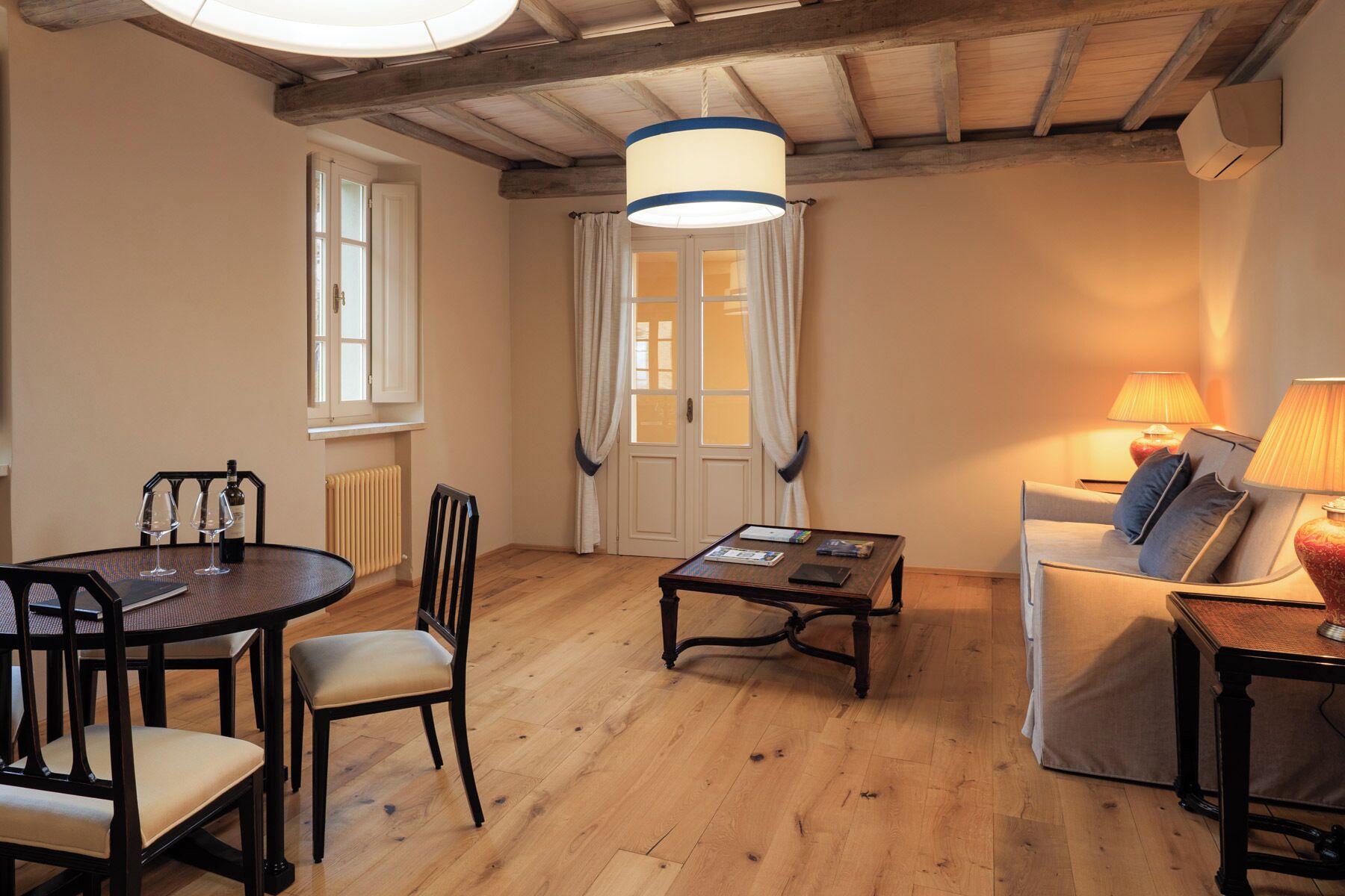 Una habitación con piso de madera, una mesa de comedor con sillas, un sofá, una lámpara y una ventana con cortinas.