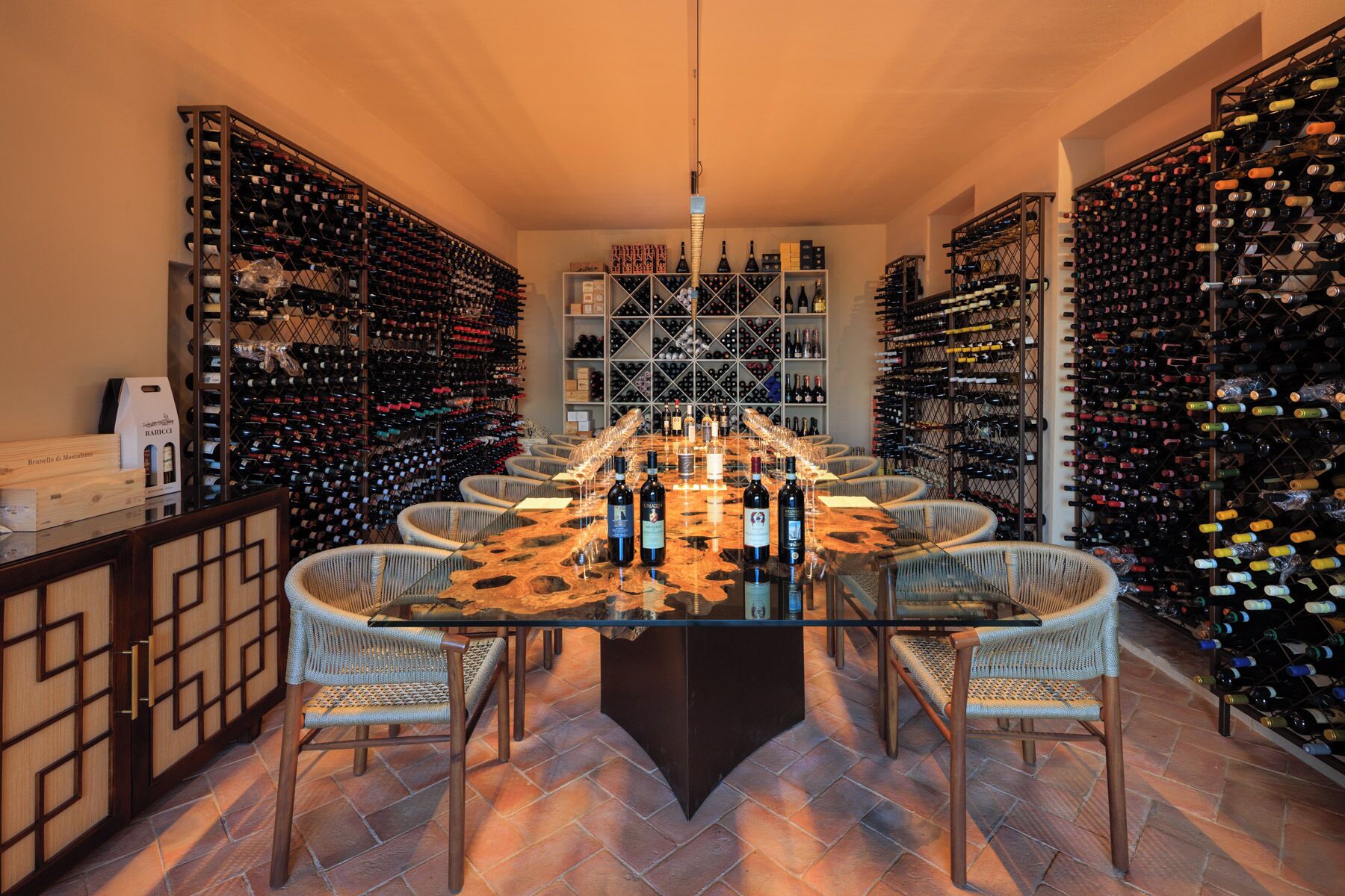 A wine cellar with a central table surrounded by wicker chairs, wine racks on both sides, and a variety of wine bottles.