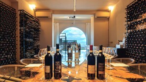 Una sala de degustación de vinos con barra de mármol, botellas de vino, copas, mobiliario de mimbre y vistas al mar a través de un amplio ventanal en arco.