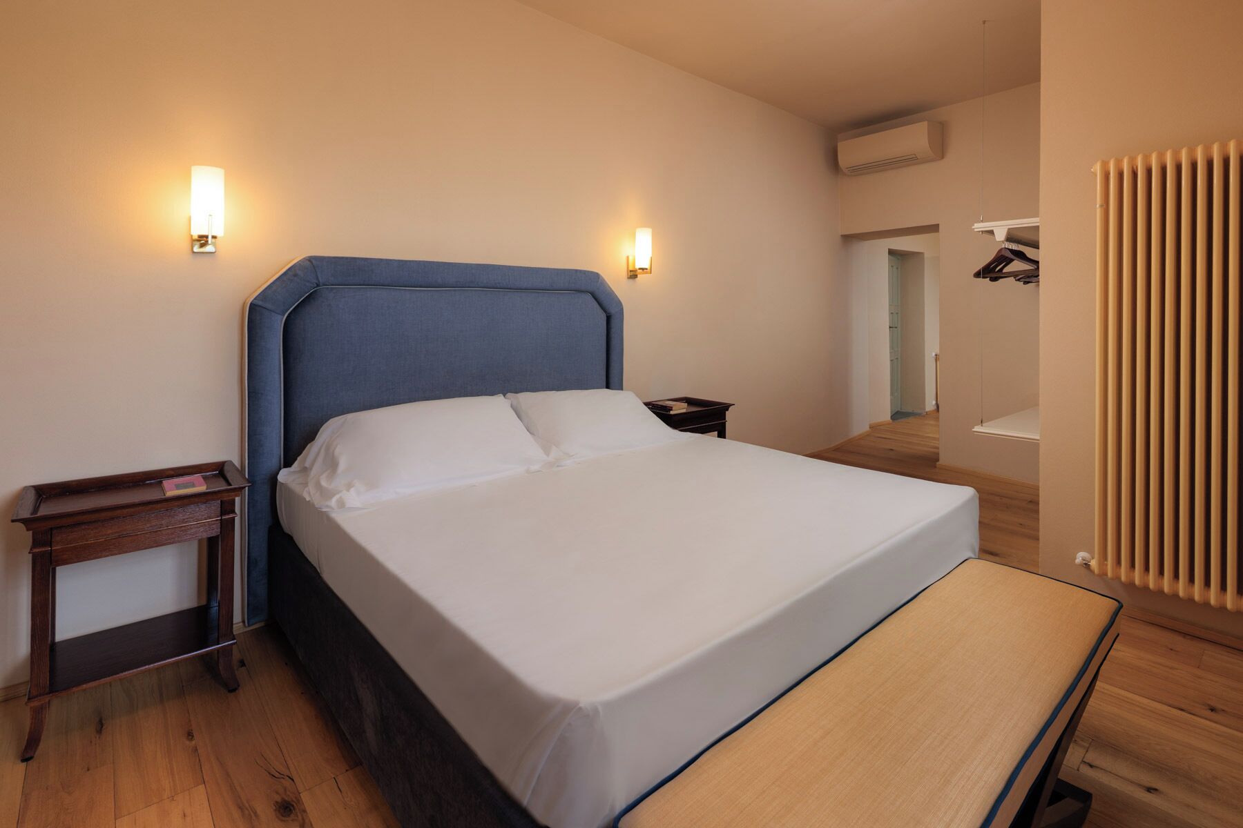 A neatly made bed in a room with a wooden floor, a small bedside table, and a wall-mounted light fixture.