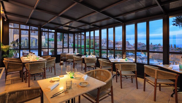 Un restaurante con mesas, sillas de mimbre, amplios ventanales y vistas a edificios y zonas verdes.
