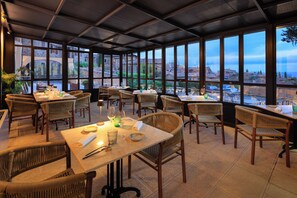 Un restaurant avec des tables et des chaises en osier, de grandes fenêtres et une vue sur les bâtiments et la verdure.