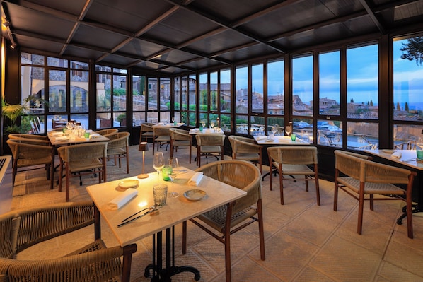 A restaurant with tables and wicker chairs, large windows, and a view of buildings and greenery.
