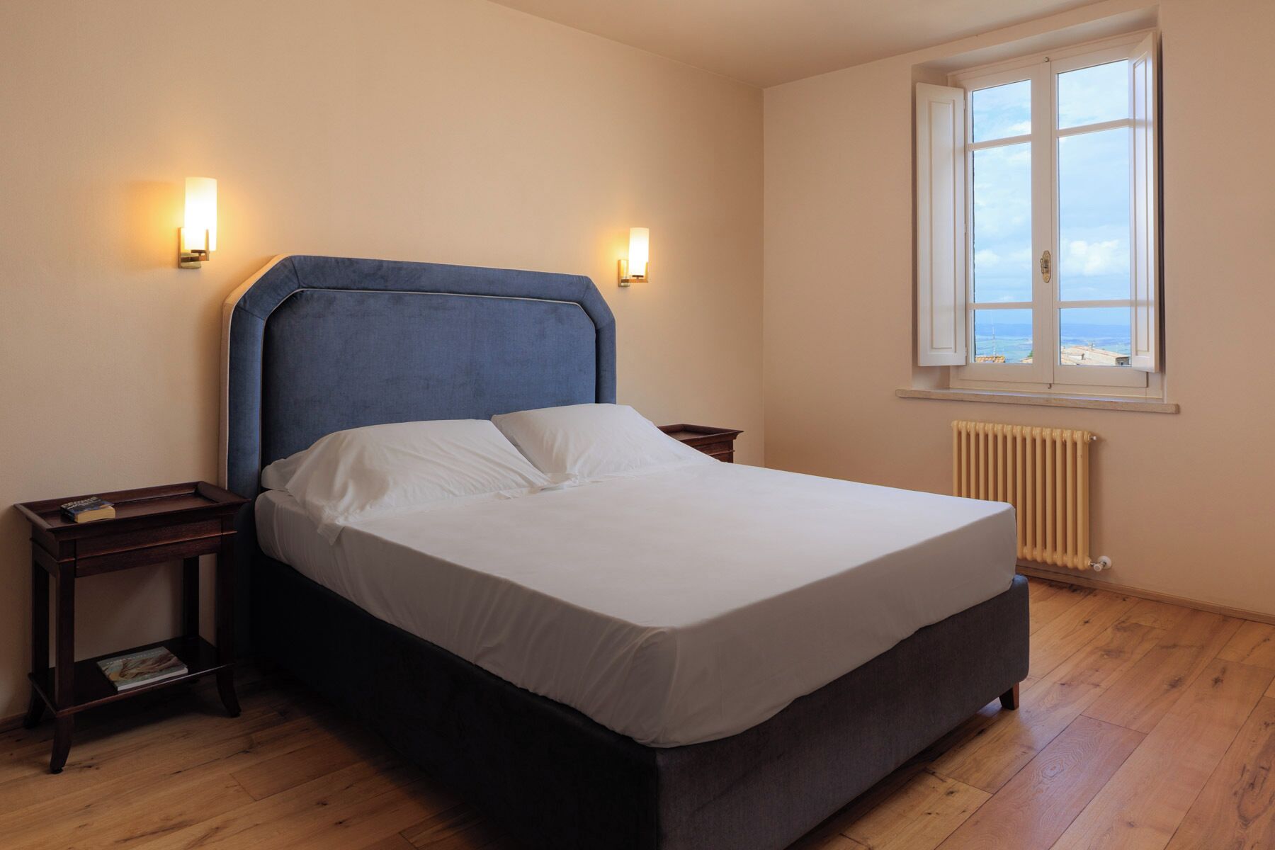 A bedroom with a bed, two bedside tables, a window with a view, and a radiator.