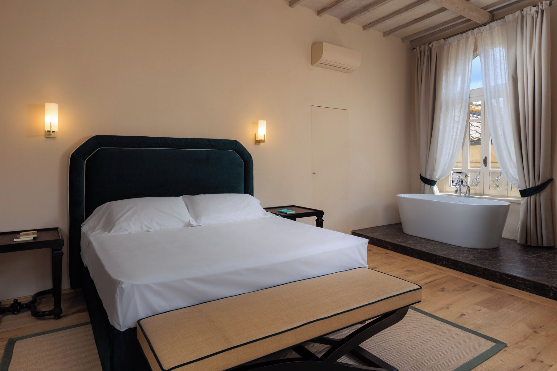 A bedroom with a bed, bedside tables, a nightstand, a window with curtains, and a bathroom with a bathtub.