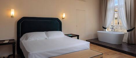 A bedroom with a bed, bedside tables, a nightstand, a window with curtains, and a bathroom with a bathtub.