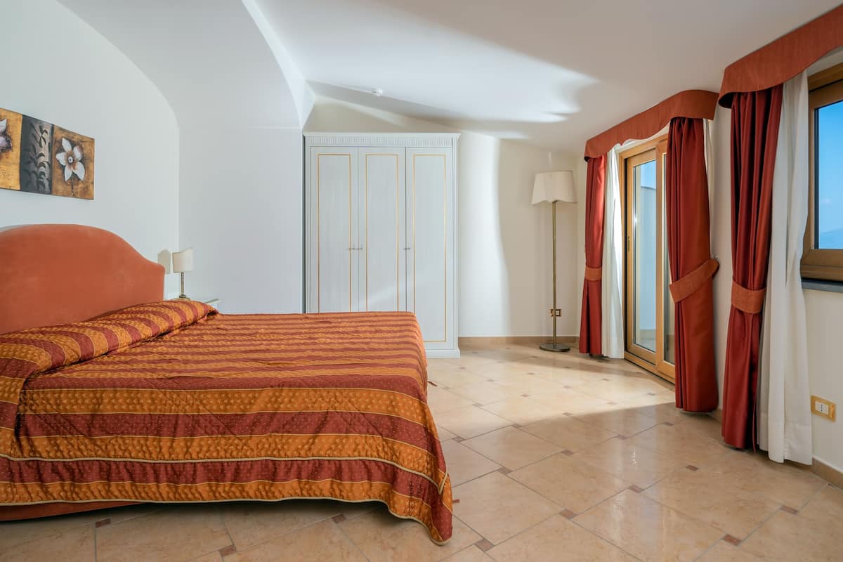 Quarto de hotel com uma cama grande, um guarda-roupa, uma porta, um candeeiro e uma janela com cortinas.