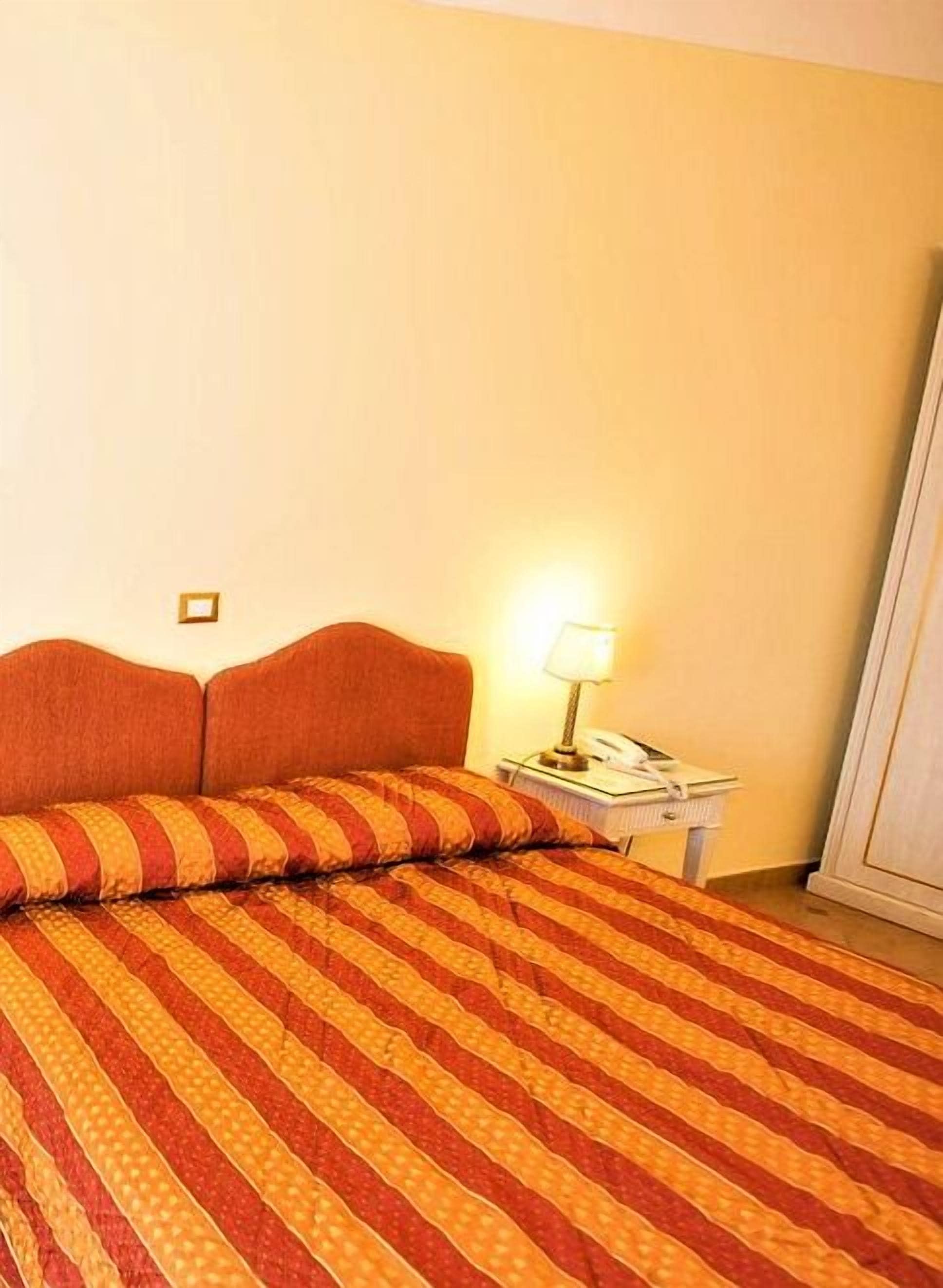 Uma cama com colcha listrada, uma mesa de cabeceira com abajur e um telefone fixado na parede.