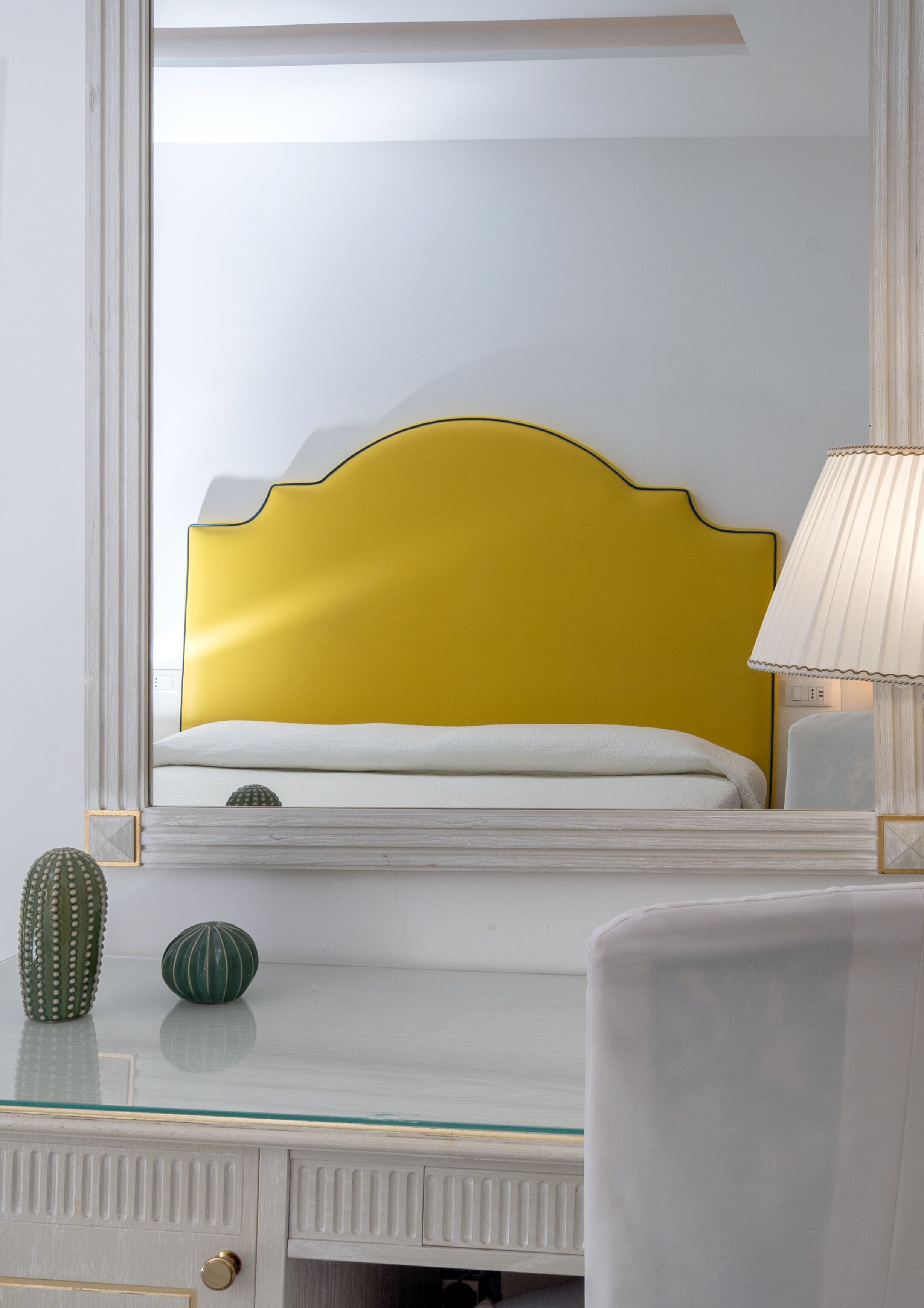 Una cama con cabecera amarilla, una lámpara de noche y jarrones decorativos sobre un tocador blanco.