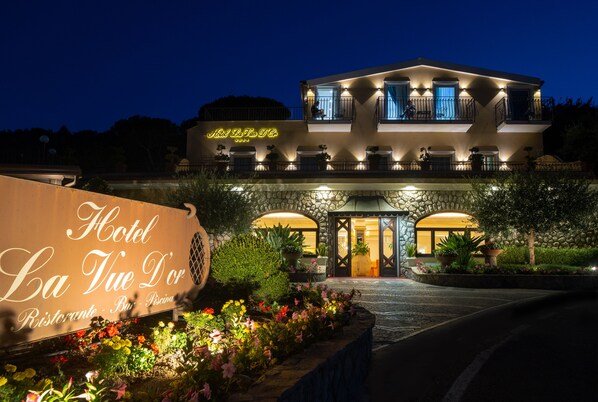 Un hotel con un letrero que dice "Hotel La Vue D'Or" y "Ristorante - Bar - Piano". Cuenta con fachada de piedra, varios balcones e iluminación nocturna.