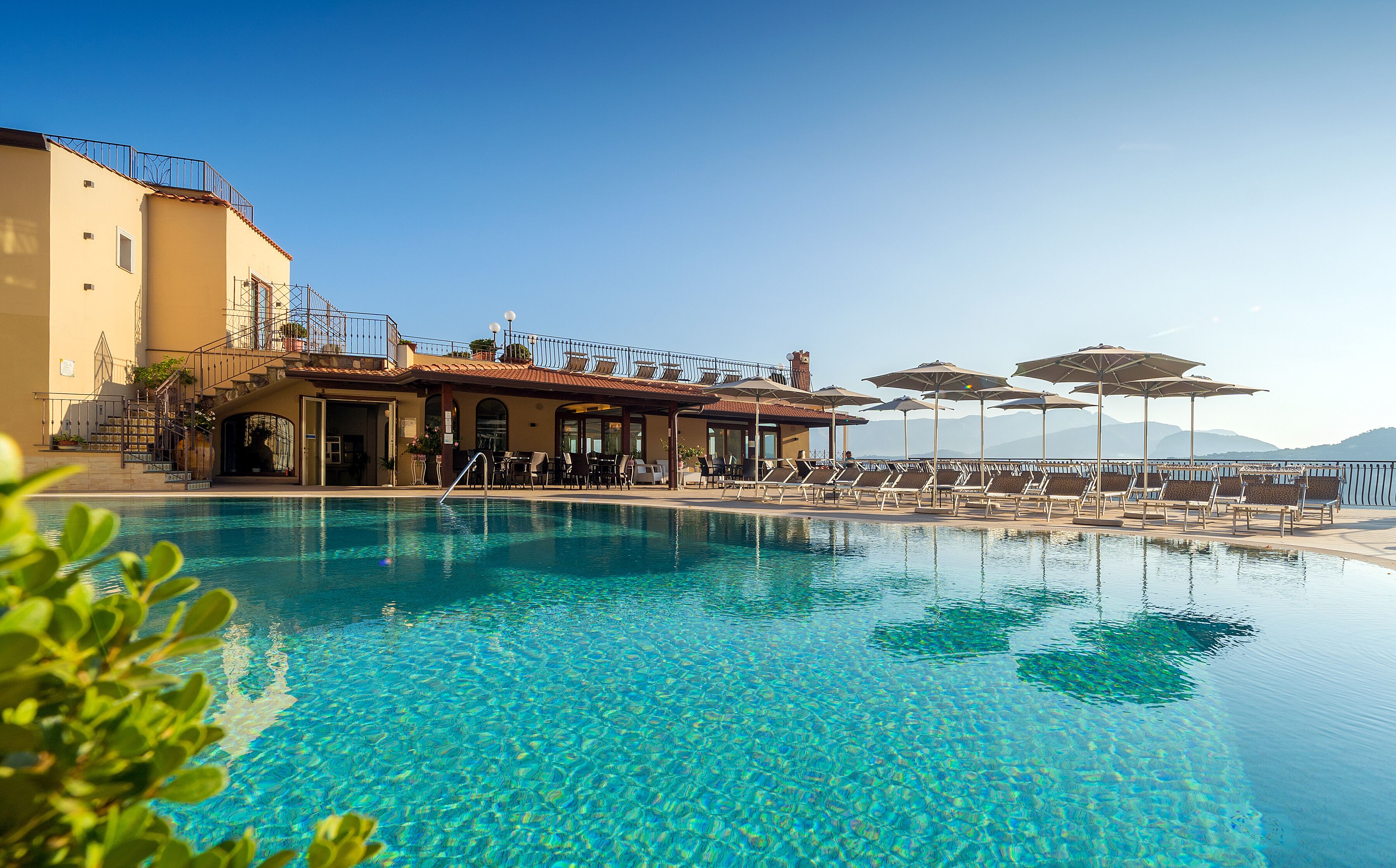 Una piscina azul cristalina con sillas de descanso y sombrillas, un edificio con balcón y montañas al fondo.