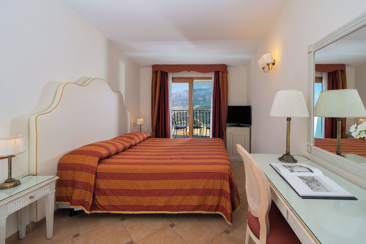 Quarto de hotel com cama, escrivaninha com luminária, TV e varanda com vista para as montanhas.