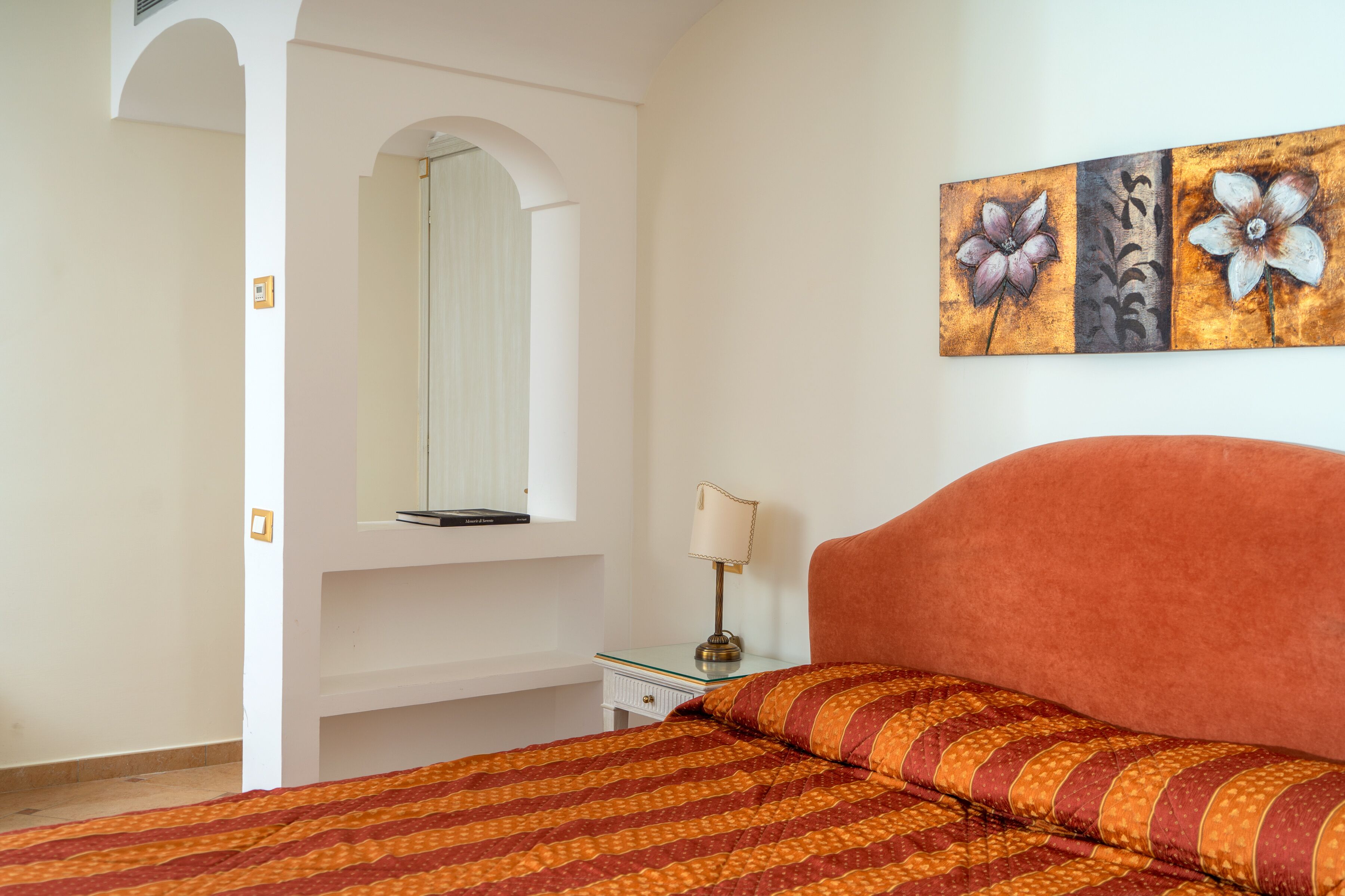 A bedroom with a bed, a nightstand, a lamp, a mirror, and a wall with a floral painting.